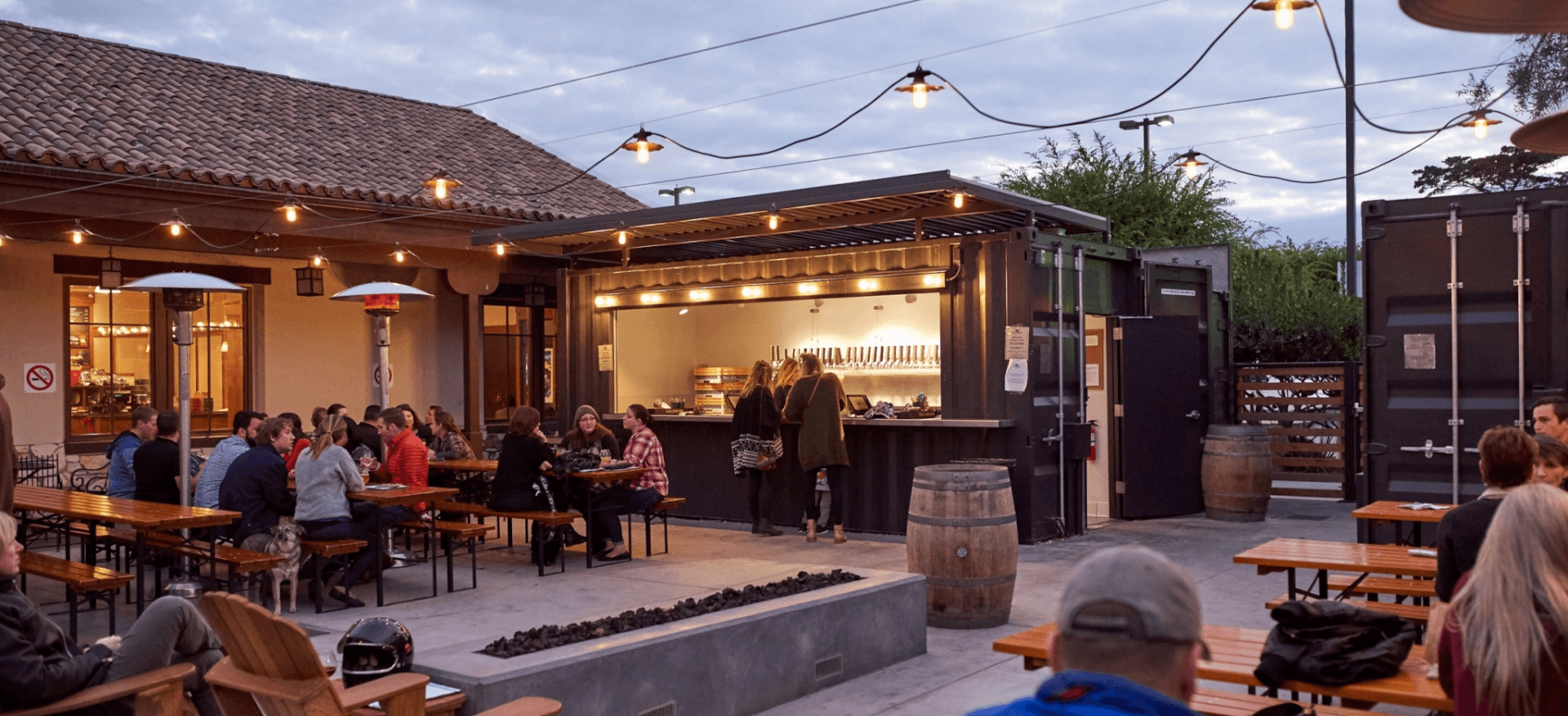 Outdoor brewery space for Fieldwork Brewing Co in Monterey, Ca. People sit in lounge chairs and picnic tables under string lights and around firepits