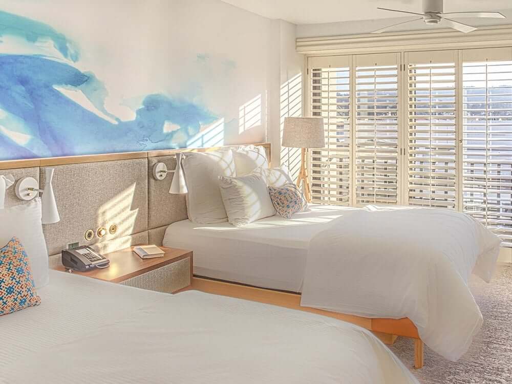 Eco-Friendly Ways to Experience Monterey Two hotel beds in room with open shutters and ocean view peaking through at the Portola Hotel & Spa in Monterey, Ca